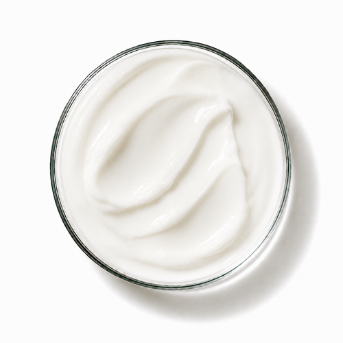 A top-down view of a clear glass jar filled with thick, creamy white lotion or cream, set against a clean white background with a soft shadow.